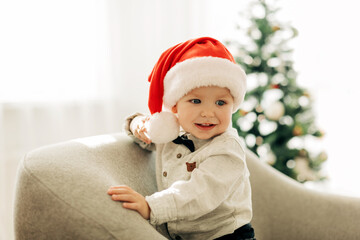 Obraz na płótnie Canvas A cheerful boy in a Santa Claus hat is sitting under a Christmas tree in an armchair and enjoying the holidays