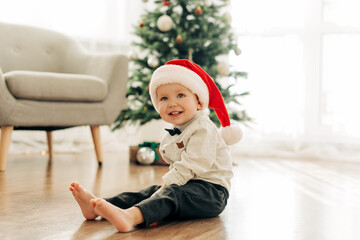 Obraz na płótnie Canvas A laughing boy in a Santa Claus hat sits on the floor against the background of a Christmas tree