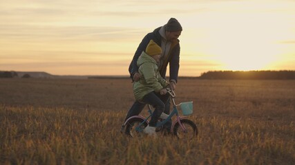 child with dad learns to ride bike at sunset, happy family, kid and dad spin pedals and wheels in light of the sun, baby learn to ride in nature, fun childhood, play on weekends with parent outdoors