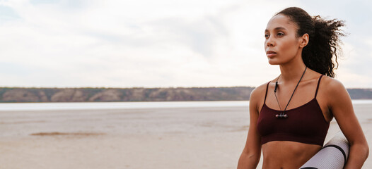 Black young sportswoman standing with fitness mat while working out
