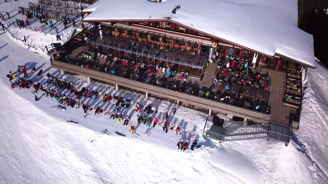 Aerial drone video of crowded ski restaurant with terrace full of people. Apres ski bar surrounded by snow with stage and live music cramped with skiers relaxing. Mountain hut overcrowded by tourists.