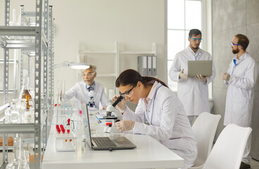 Fototapeta premium Clinic lab examination. Young smart woman examines test samples with a microscope in a modern medical laboratory. Concept of science, chemistry, technology, biology and people.