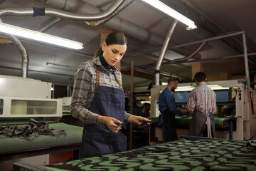 Portrait of young woman working in workshop of footwear factory. Serious female worker standing at cutting machine and looking at cut details of leather material. Concept of shoe manufacturing process