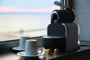 Coffee machine with capsules and two cups ready to be used near by a window.