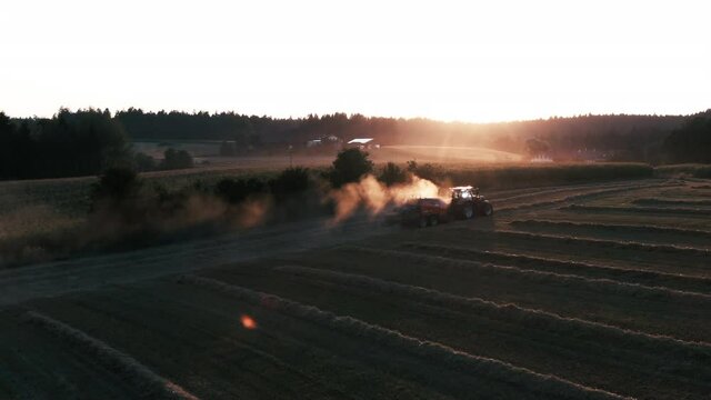 Aerial drone shot of a tractor with a harvesting machines creating blocky hay balls and the sunset in the background with beautiful backlight and a lot of dust.