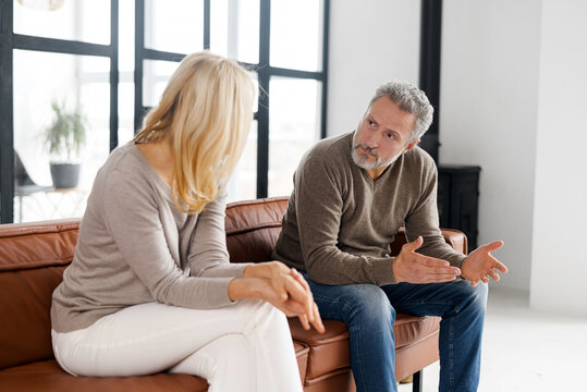 Frustrated, Depressed And Unhappy Crying Middle-aged Woman Sits On The Foreground, Mature Grey-haired Man In Out Of Focus On The Background. Matured Couple Is Close To Divorce