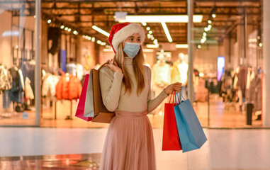 A young girl model in a Santa hat, a protective medical mask and with shopping bags is shopping for gifts in the mall.Shopper. Sales. Shopping Center. Consumerism and people new normal concept.
