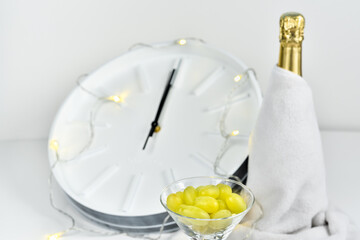 Grapes in a glass against the background of a blurry clock and a bottle of champagne. Spanish tradition of eating twelve grapes to celebrate New Year.