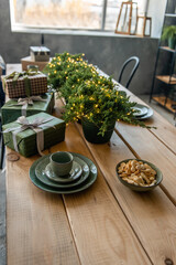 On a wooden table there are festive green dishes, wrapped boxes of gifts, thuja in pots with a garland, children's treats. Christmas holiday atmosphere. Table setting for dinner . Copy space