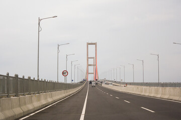 Fototapeta na wymiar Scene of the famous Suramadu Bridge and its red suspension steel cables with cars and lamp post on road and cloudy sky background. 