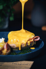 egg Benedict with hollandaise sauce on dark blue plate on table in kitchen