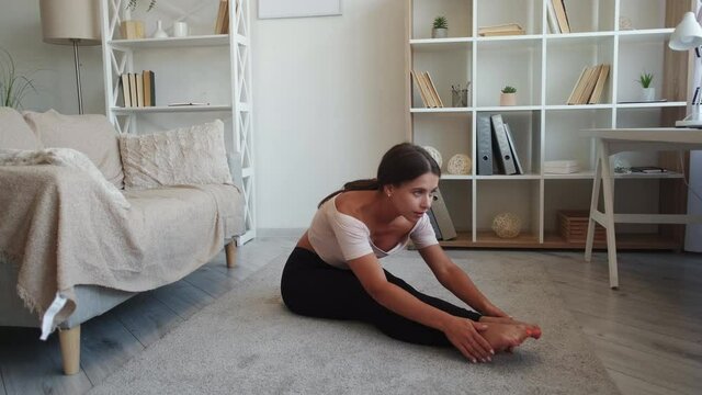 Home gymnastic. Slim woman. Fit training. Pretty lady doing stretching workout bending forward in light living-room interior.