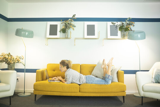 Modern woman use laptop computer laying on the sofa in stylish living room at home or hotel hall. Concept of female people and smart working job activity relaxed on the yellow couch