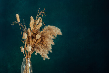 Bouquet of beige dried flowers in a glass vase on green-blue background.