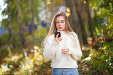 beautiful autumnal young girl in a white sweater in park with smartphone among the trees, caucasian girl with beautiful long hair and full lips, portrait of a girl soft focus and beautiful bokeh