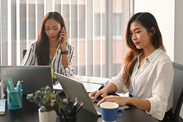 Photo of two office women working together at the modern working desk surrounded by a computer laptop and office equipment.