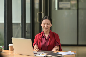 Photo of a beautiful businesswoman smiling and sitting with crossed arms at the wooden working desk surrounded by a computer laptop and office equipment.