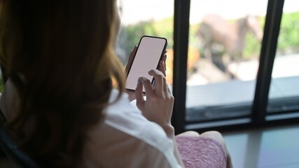 Behind view of a young woman using a white blank screen smartphone in the comfortable living room as a background.