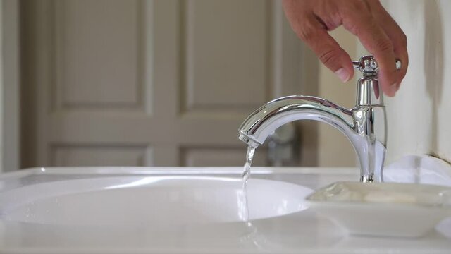 Man turn valve and shut off water pouring from faucet to bathroom sink, slow motion shot. Few drops trickle down after man move out his hand. Close up view of spigot at typical washroom, blurred back