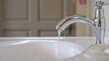 A thin trickle of water pours from the faucet in sink at bathroom, man closes the tap and stop the leakage. Close up slow motion shot of polished spigot, blurred door on background