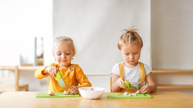 Cute kids toddlers girls at the table cut apples with safe knives, children cut food themselves, montessori and independence in kindergarten