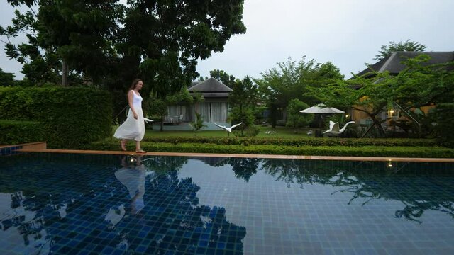 Woman wearing white clothes walk on edge of swimming pool, camera turn right to follow her motion. Tourist lady rest at resort garden in late hour, dim natural light