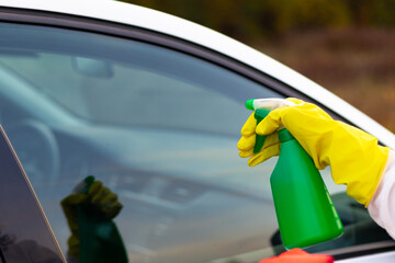 A hand in a yellow rubber glove sprays water from a spray bottle onto a car window on a warm autumn day. Wet cleaning. Selective focus. Close-up