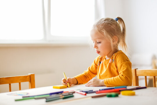 Cute sunny girl child blonde with a ponytail at the table draws with multicolored felt-tip pens, montessori and creativity, creative development in kindergarten