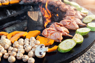 Close-up detail view of many tasty raw marinated quails and vegetables and mushrooms grilled cooked at round steel iron firepit hearth table surface brazier with burning firewoods. Barbecue yard home