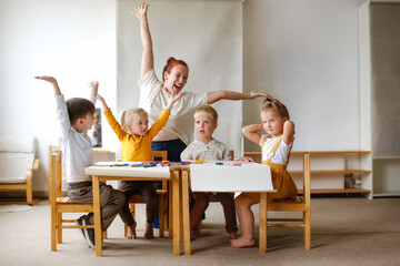 cheerful teacher woman with a group of cute preschool children at the table draw on paper with...