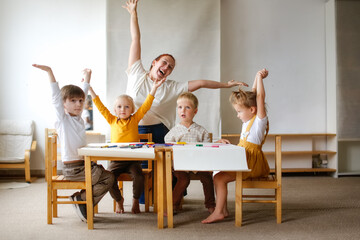 cheerful teacher woman with a group of cute preschool children at the table draw on paper with...