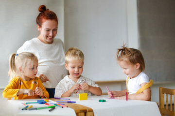 cheerful teacher woman with a group of cute preschool children at the table draw on paper with multicolored felt-tip pens, montessori and creativity, creative development and lesson in kindergarten
