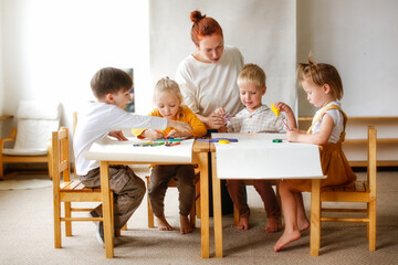 cheerful teacher woman with a group of cute preschool children at the table draw on paper with multicolored felt-tip pens, montessori and creativity, creative development and lesson in kindergarten - obrazy, fototapety, plakaty