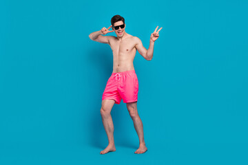 Photo of crazy carefree clubber man show v-sign wear sunglass swimwear shorts isolated blue color background