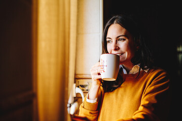 Portrait of a content young woman with coffee cup looking out through cabin window