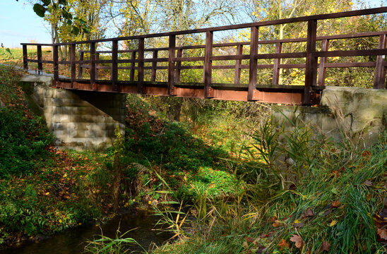 pedestrian bridge over a small gorge and a stream. it consists of two steel crossbeams. as the surface and railing of the bridge is made of brown painted planks. is long and narrow. in the wild