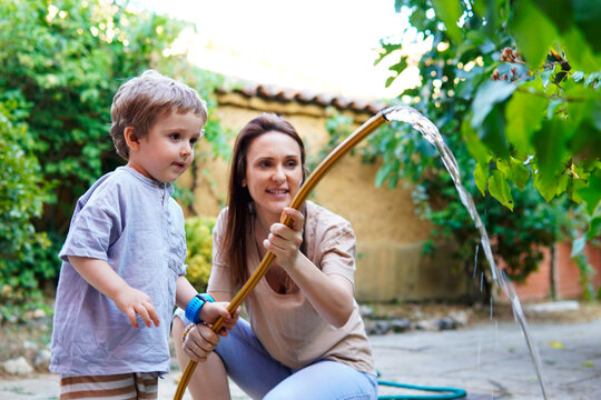 young woman beautiful middle-aged mother with her three-year-old son watering with a hose in the garden at home. family time