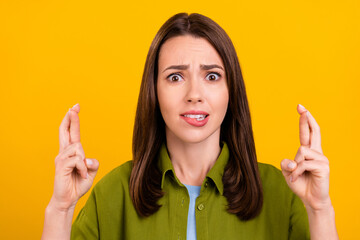 Photo of unsure unhappy young woman dressed green shirt biting lip fingers crossed isolated yellow color background