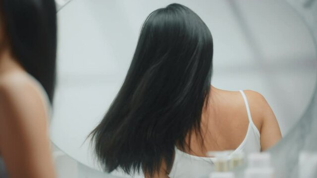 Beautiful Asian Woman Turns Around Her Lush Black Hair Flying. Young Adult Female Admires Herself in Bathroom Mirror. Concept for Happiness, Wellbeing, Natural Beauty, Organic Skincare Products