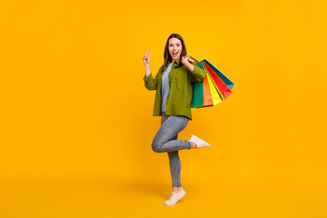 Photo of sweet excited young woman dressed green shirt holding bargains showing v-sign smiling isolated yellow color background
