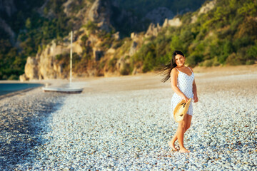 Fototapeta premium A cute girl in a light summer sundress and a straw hat in her hands is walking along a deserted paradise beach. Turkey, Kemer, Cirali.
