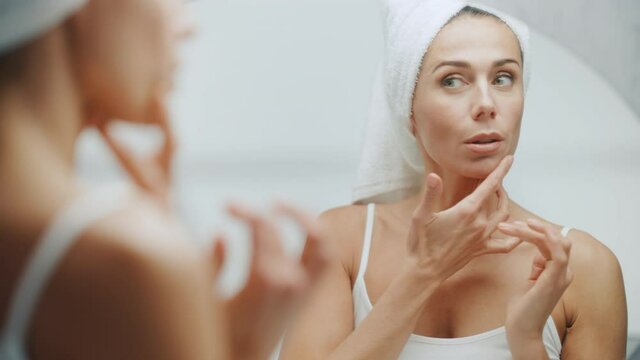 Beautiful Caucasian Woman Touches Her Perfect Face, Enjoying Her Ideal Wrinkle Free Skin, Sensually Smiles into Bathroom Mirror. Happy Middle Aged Female Uses Natural Cosmetic Skincare Products