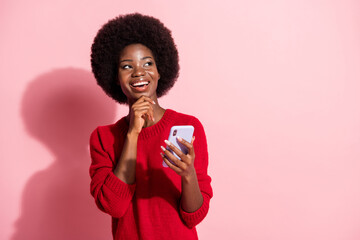 Photo of minded happy dark skin woman look empty space hold hand chin phone isolated on pink color...