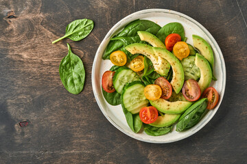 Avocado, red, yellow, black cherry tomato, spinach and cucumber fresh salad with spices pepper and olive oil in grey bowl old wooden table background. Healthy food concept. Top view.
