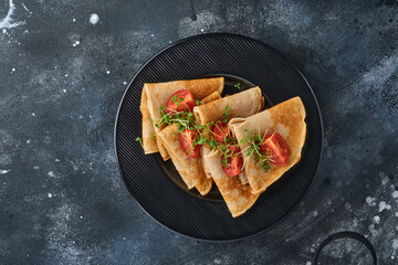 Stack of gluten free buckwheat flour crepes pancakes with cherry tomatoes and arugula microgreens on black plate on dark background, homemade healthy baking for breakfast. Mock up. Copy space.