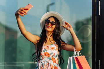 woman, holding bags with purchases , and taking a selfie on a smartphone while standing near a shopping center