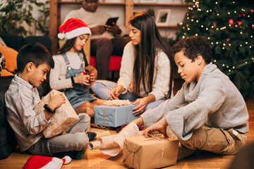 Happy mother sitting with her children in front of the Christmas tree at Christmas and unwrapping presents. Multicultural family celebrating Christmas.