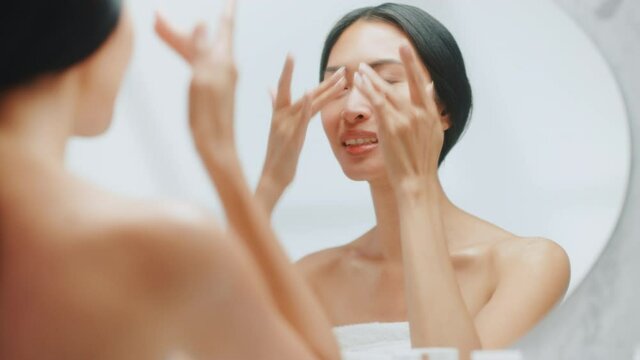 Portrait of Beautiful Asian Woman Gently Applying Face Cream Looking in Bathroom Mirror. Young Adult Female Makes Her Skin Soft, Smooth with Natural Moisturizing Cosmetics Skincare Product