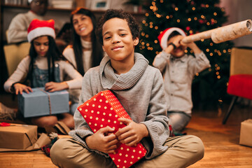 A happy mixed-race boy sitting on the floor at home at Christmas and holding a gift in his hands....