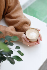 a cup with cappuccino in the hands of a woman on a white table